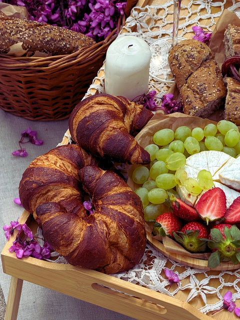 Picnic tray with golden croissants, green grapes, brie cheese, strawberries, and rustic bread on a lace tablecloth.