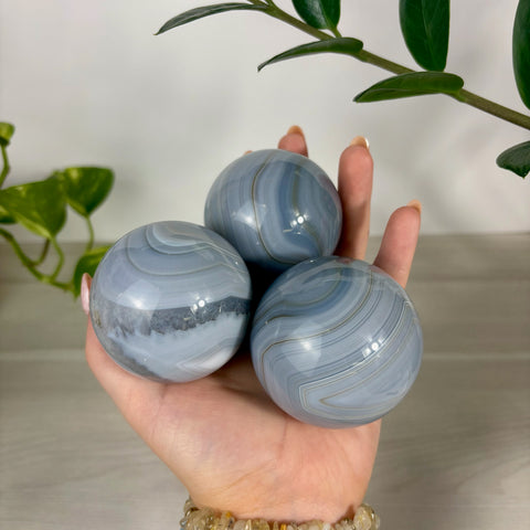 Three polished blue banded agate spheres with natural grey swirls held in a hand against a white background.