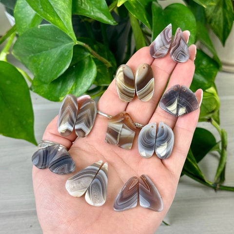 Botswana Agate cabochon pairs in various shapes and natural brown, grey, and white bands, held in a hand.