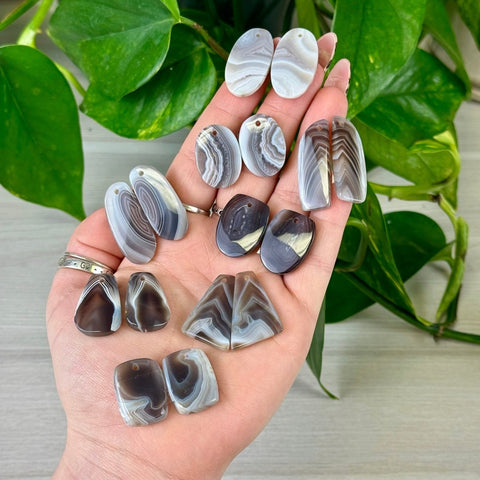 Hand holding various Botswana Agate cabochon pairs with predrilled holes, featuring gray, white, and brown bands.
