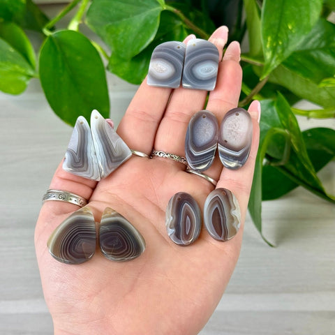 Botswana Agate cabochon pairs in gray and white, various shapes, with predrilled holes, held in a hand.