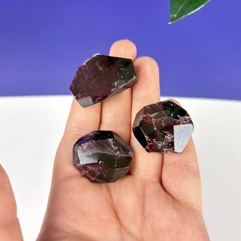 Three polished, deep red garnet Cherry Blood Gusher crystals held in a palm against a blue and white background.