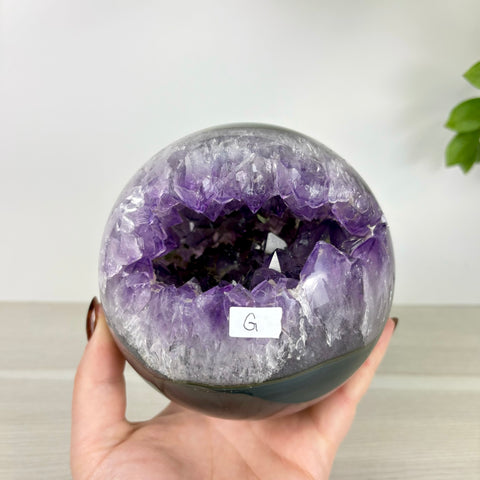 Polished amethyst geode sphere with deep purple crystals held in a hand, labeled with a small white tag marked G.