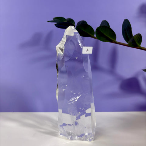 Clear quartz polyhedron crystal with faceted edges, labeled A, standing on a white surface against a purple background.