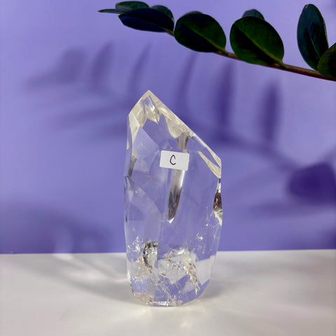 Clear quartz polyhedron crystal with faceted edges and internal inclusions, labeled with a white C sticker on a purple background.
