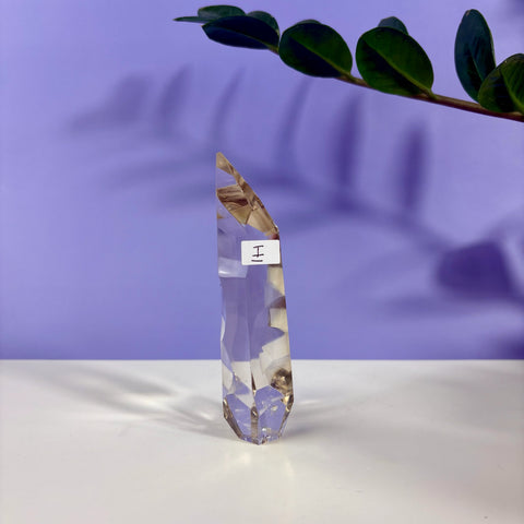Clear quartz polyhedron crystal point with geometric facets, labeled I, standing on a white surface against a purple background.