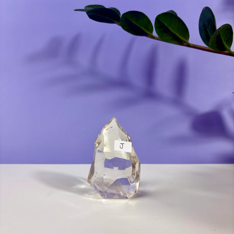 Clear quartz polyhedron crystal labeled J, featuring a faceted, transparent design on a white surface against a purple background.