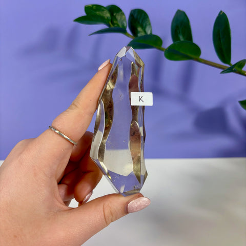 Hand holding a faceted, transparent clear quartz polyhedron crystal labeled with a K sticker against a purple background.