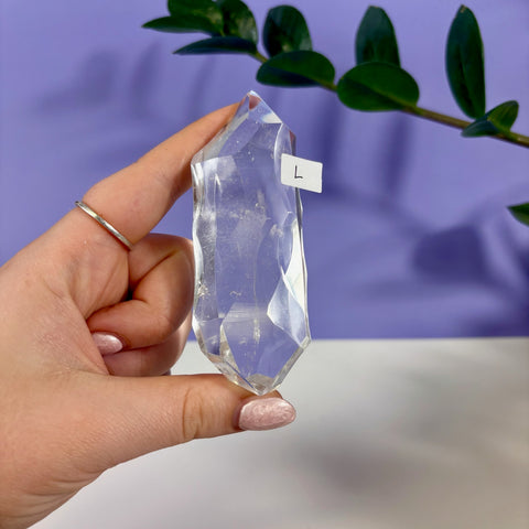 Hand holding a large, transparent clear quartz polyhedron crystal with geometric facets against a purple background.