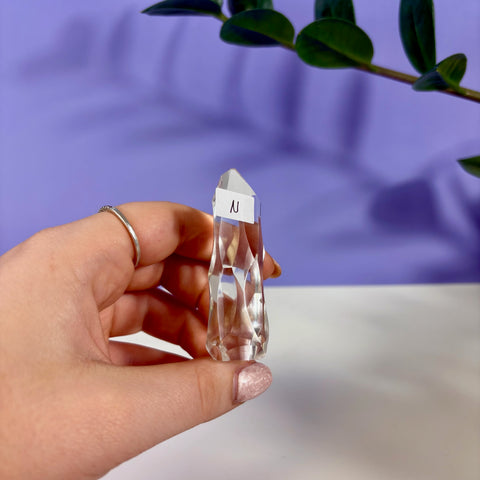 Hand holding a clear quartz polyhedron crystal with faceted edges against a purple background.