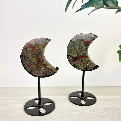 Two crescent moon-shaped Dragon's Blood jasper stones with red and green patterns, displayed on black metal stands.