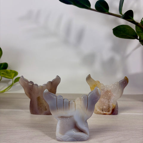 Three polished druzy agate mermaid tail carvings in shades of grey, tan, and white on a wooden surface.
