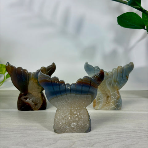 Three polished druzy agate mermaid tail carvings in blue, brown, and grey tones displayed on a white wooden surface.