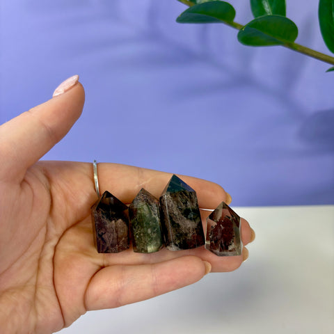 Four small Garden Quartz towers with mossy green and brown inclusions held in a palm against a purple background.