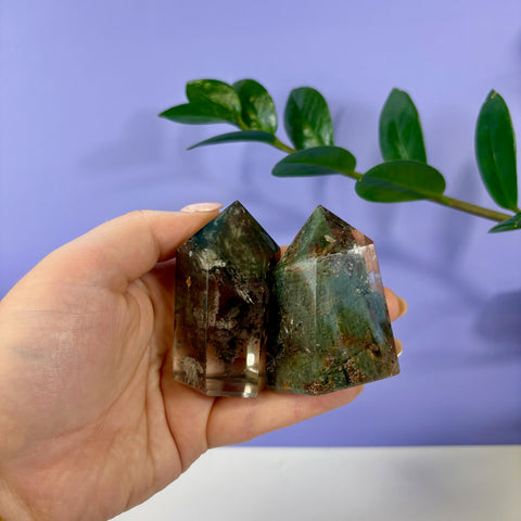 Two polished Garden Quartz towers with green moss-like inclusions held in a hand against a purple background.
