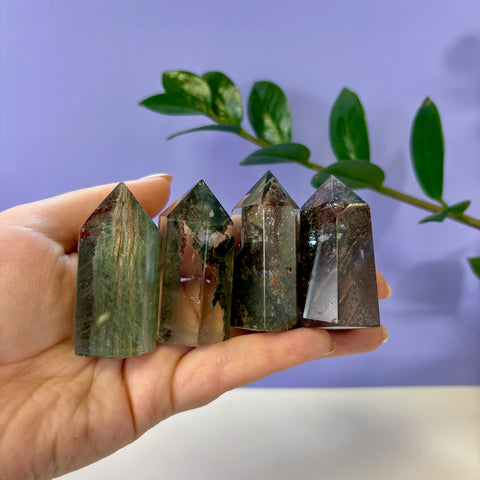 Four polished Garden Quartz towers with green and brown mineral inclusions held in a hand against a purple background.