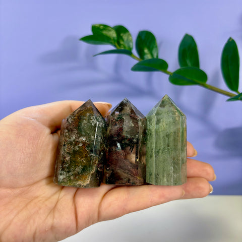 Three polished Garden Quartz towers with green and earthy mineral inclusions held in a hand against a purple background.