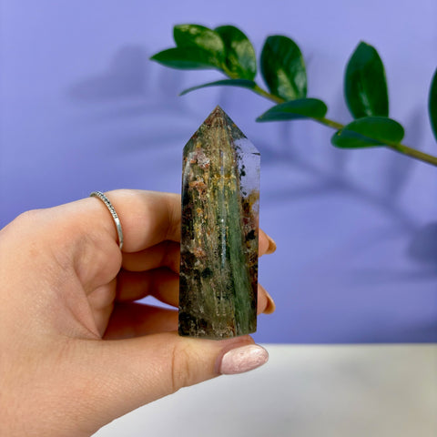 Hand holding a clear Garden Quartz tower with green and earthy mineral inclusions against a purple background.