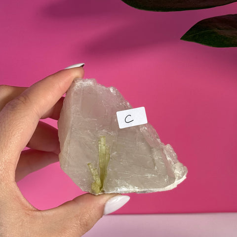 Hand holding a raw, light green tourmaline specimen (variant C) against a pink background.