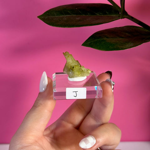 Vibrant green tourmaline specimen on a clear acrylic stand, held by a hand with white nails.