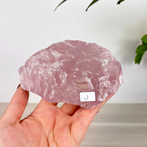 Large, raw chunk of translucent pale pink rose quartz held in a hand, featuring a small white label with the letter J.