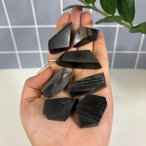 Hand holding six small, polished black Hypersthene free form crystals with metallic, shimmering linear patterns.