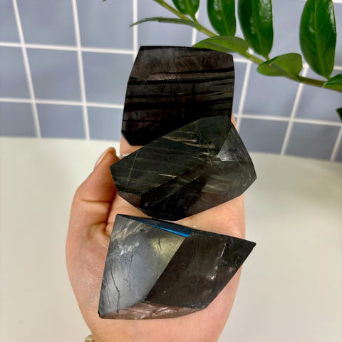 Three polished Hypersthene free form crystals with dark, metallic sheen held in a hand against a tiled background.