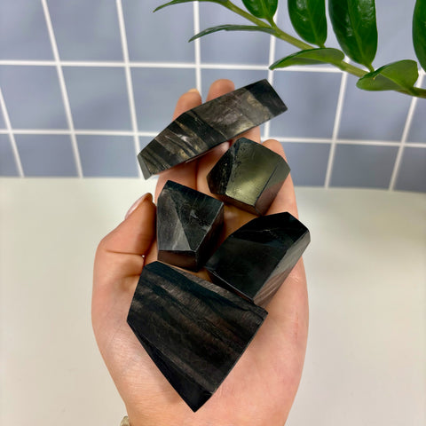 Hand holding five polished Hypersthene free form crystals with dark, metallic, and striped patterns.