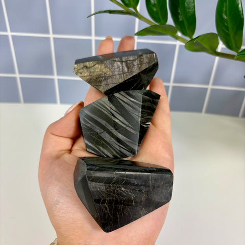 Three polished Hypersthene free form crystals with dark, metallic striped patterns held in a hand.