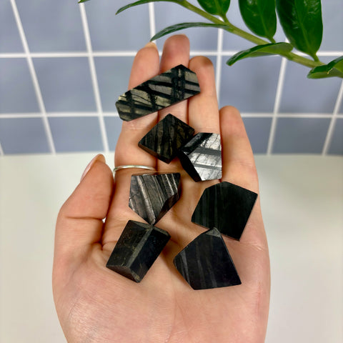 Hand holding several small, polished black Hypersthene free form stones with metallic, shimmering linear patterns.