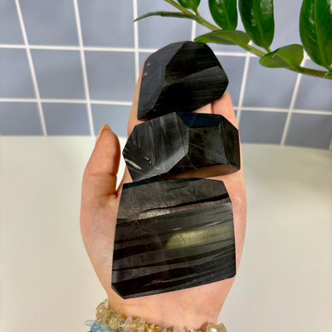 Three polished black hypersthene free form stones with metallic sheen held in a palm against a tiled background.