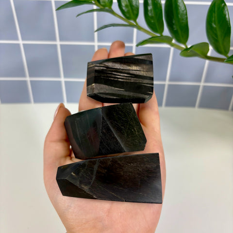Three polished black hypersthene free form crystals held in a hand against a tiled background.