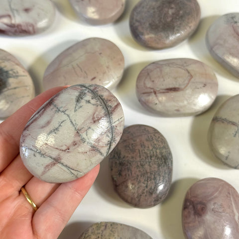 Smooth, oval pink and grey imperfect painting stone palm stone held in hand, surrounded by similar stones.