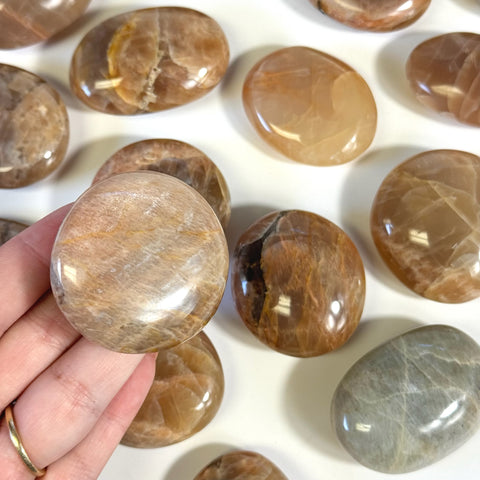 Hand holding an imperfect peach moonstone palm stone, surrounded by similar polished, oval-shaped stones.