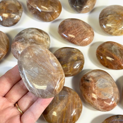 Hand holding a polished, imperfect peach moonstone palm stone with brown and orange tones.