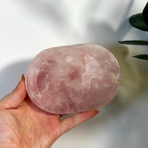 Hand holding a polished, oval, imperfect rose quartz plate with soft pink hues.