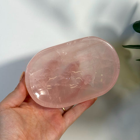 Hand holding a polished, oval imperfect rose quartz plate with natural pink hues.
