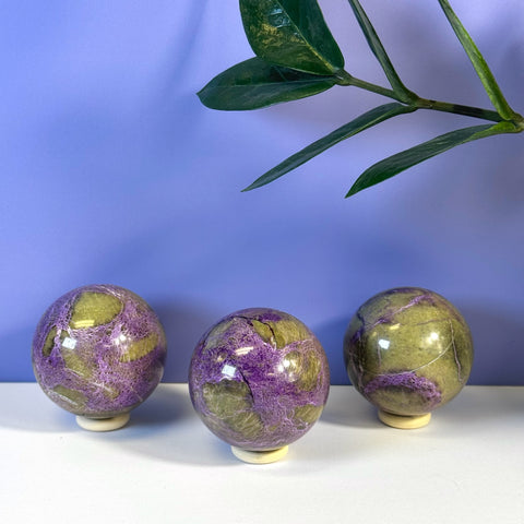 Three imperfect stichtite spheres with vibrant purple and green marbling on white stands.