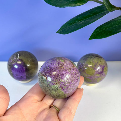 Hand holding a vibrant purple and green imperfect stichtite sphere. Two more spheres are in the background.