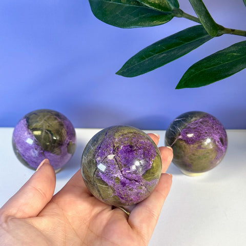 Hand holding a polished, imperfect stichtite sphere with vibrant purple and olive green colors.