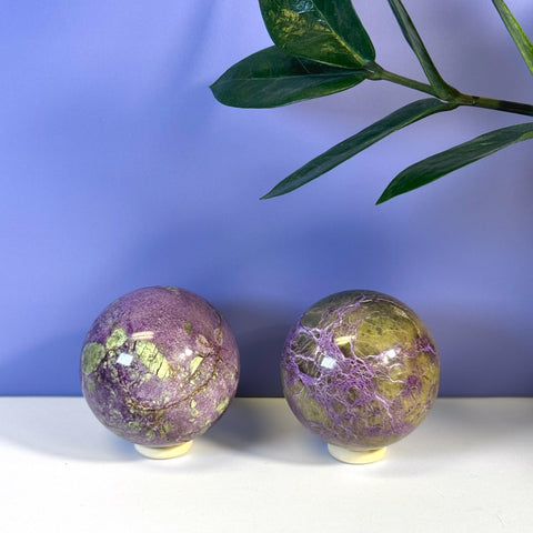 Two imperfect stichtite spheres, one purple with green, one green with purple, on white stands.