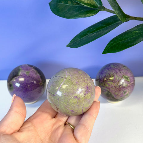 Hand holding an imperfect stichtite sphere with purple and green hues, showing natural cracks.