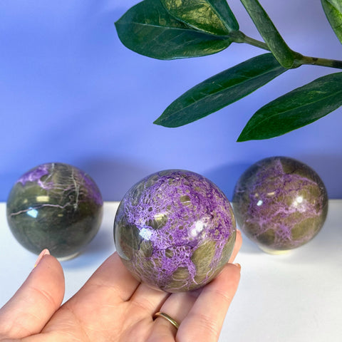 Hand holding an imperfect stichtite sphere with vibrant purple and dark green patterns.