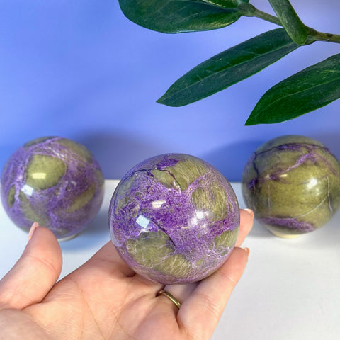 Hand holding a polished, imperfect stichtite sphere with vibrant purple and olive green patterns.