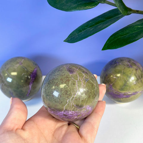 Green and purple imperfect stichtite sphere held in hand, with two more in background.
