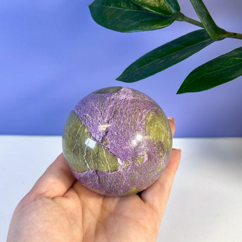Hand holding an imperfect stichtite sphere with vibrant purple and olive green patterns.