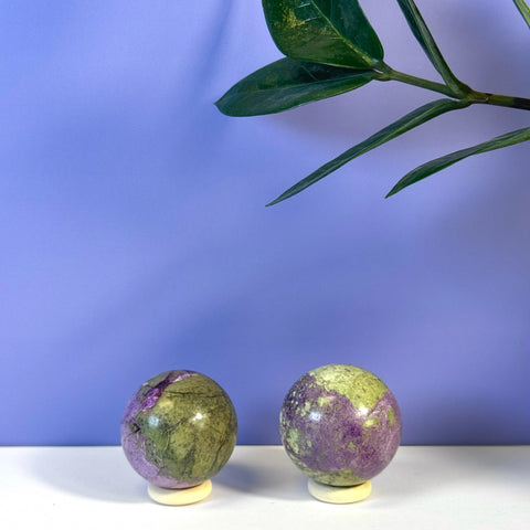 Two imperfect stichtite spheres, green and purple, on white stands against a lavender background.