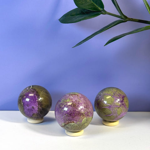 Three imperfect stichtite spheres with vibrant purple and olive green patterns on white stands.