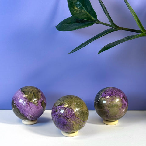 Three imperfect stichtite spheres, purple and olive green, on white stands against a purple wall.