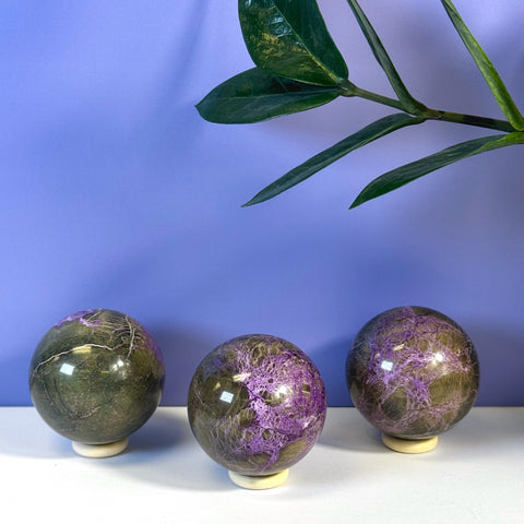 Three imperfect stichtite spheres with purple and green patterns on white stands.